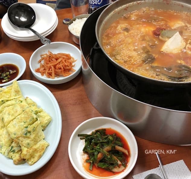 A bubbling pot of Gwanghwamun-jip kimchi-jjigae on a tabletop burner beside a plate of sliced egg roll and Korean side dishes