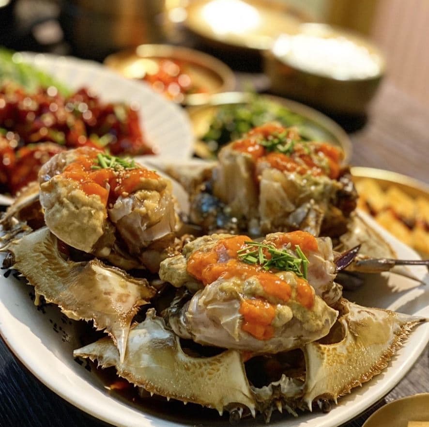 A plate of Keungiwajip ganjang-gejang — soy sauce–marinated raw crabs topped with orange roe and chopped chives, with banchan behind