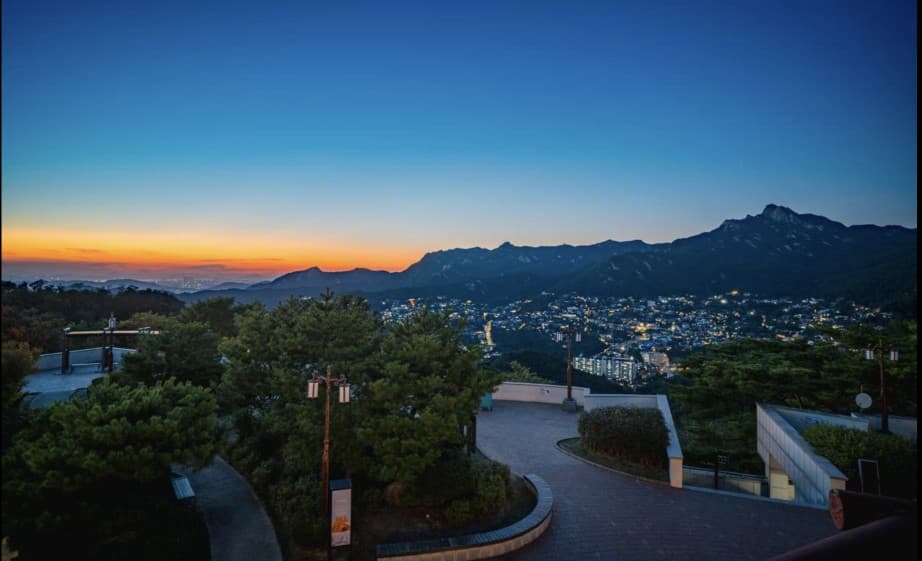 The curved brick plaza and pine trees at the Bukak Skyway Palgakjeong overlook, looking over city lights and mountain ridges at dusk