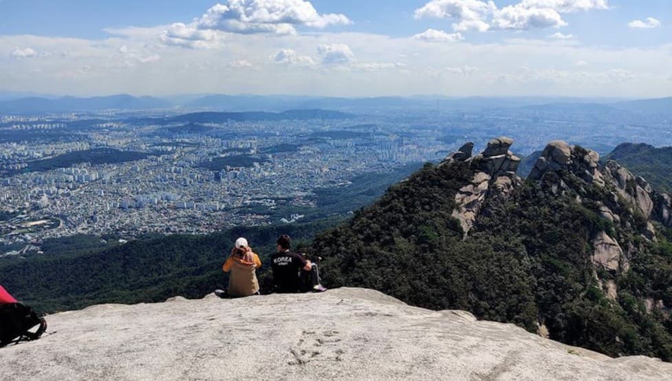 Two hikers resting on a rocky summit overlooking the sweeping Seoul cityscape under a blue sky