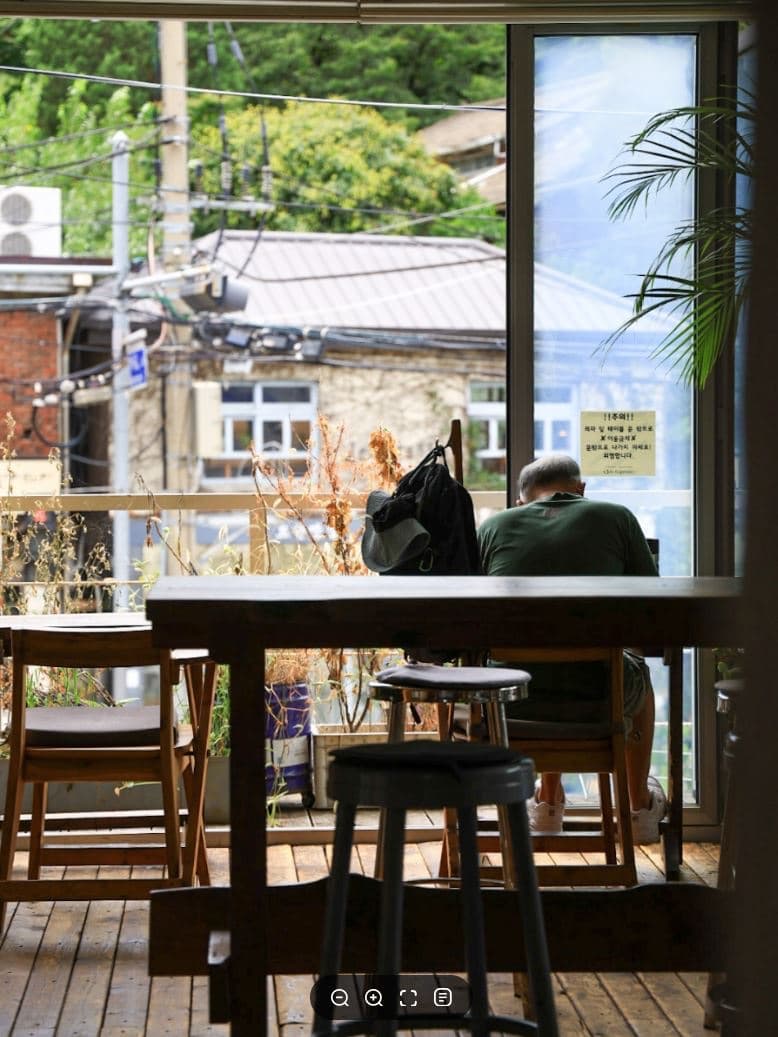 A cozy Buam-dong cafe terrace with wooden tables and stools looking out a large window onto green hills and old houses