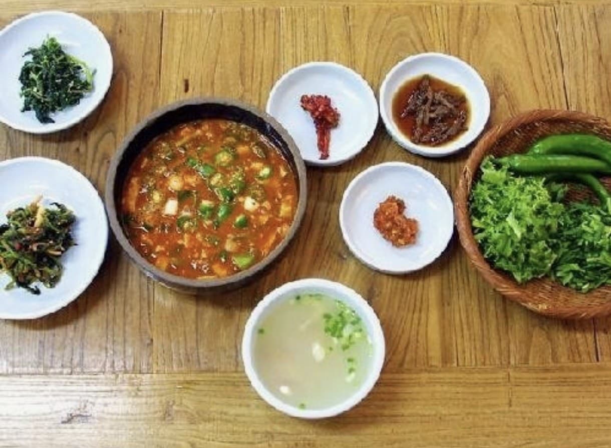 A rustic doenjang-jjigae set — bubbling soybean-paste stew with seasoned greens and ssam vegetables in a wicker basket