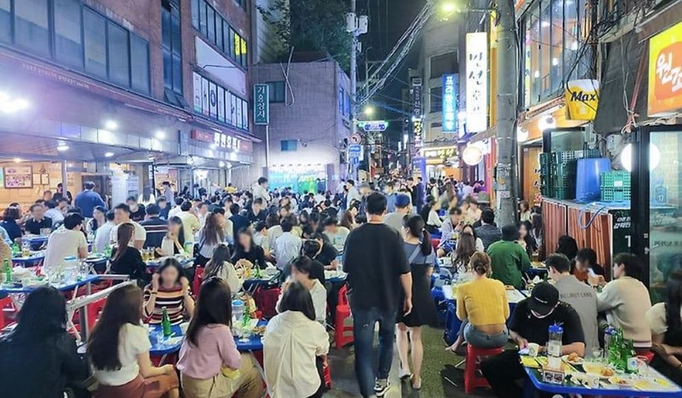 A packed nighttime Euljiro pojangmacha street with crowds seated at red and blue plastic tables and stools eating and drinking beneath neon signs and string lights