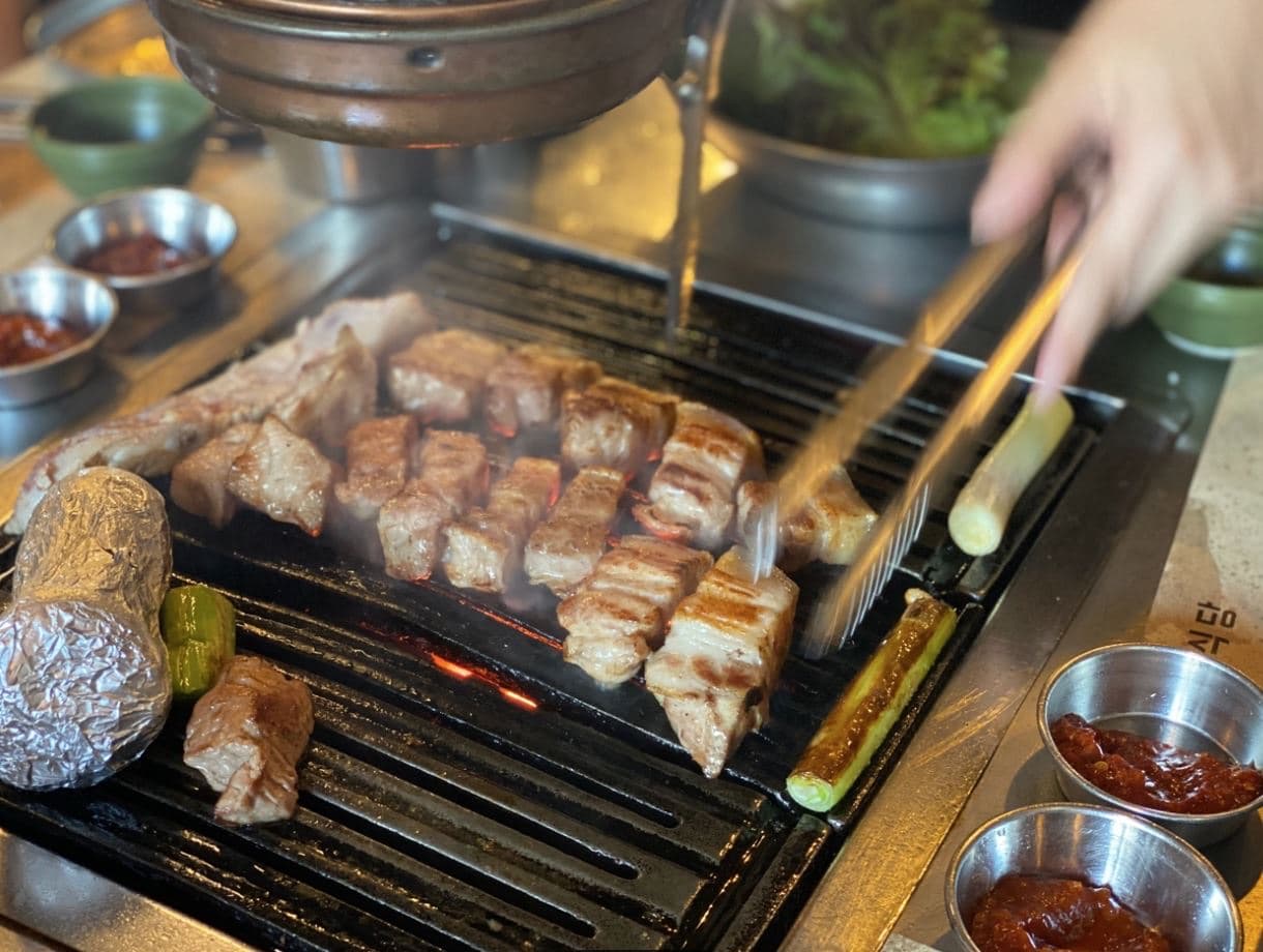 Thick cuts of pork belly grilling over charcoal on a black cast-iron grill with rising steam, foil-wrapped garlic, green chilies, and bowls of ssamjang