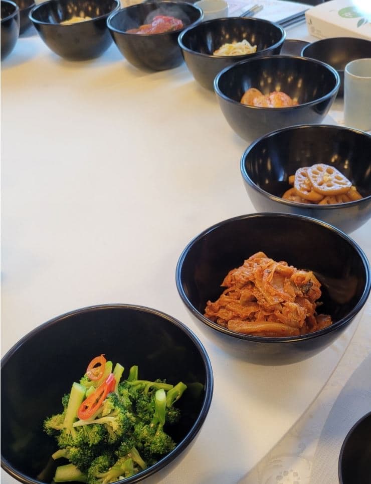 Close-up of black bowls of colorful Korean Buddhist temple banchan — seasoned broccoli with red chili, spicy braised kimchi and mushrooms, and glazed lotus root