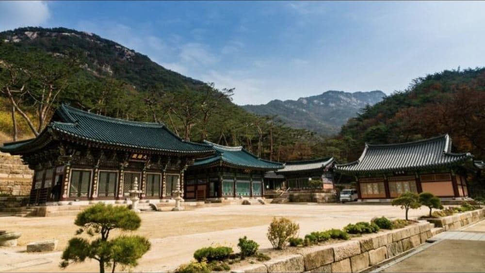 Jinkwansa Temple's traditional Korean hanok halls with green-tiled roofs at the foot of a forested mountain under a clear blue sky