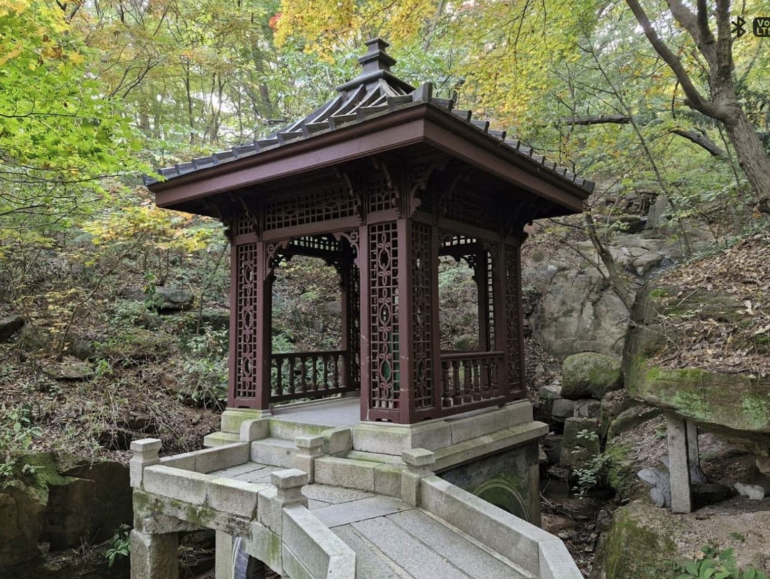An ornate dark-wood pavilion with lattice panels standing on a stone bridge amid a wooded gorge at Seokpajeong